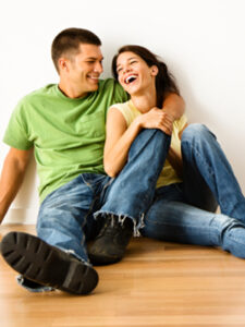 Attractive young adult couple sitting close on hardwood floor in home smiling and laughing.